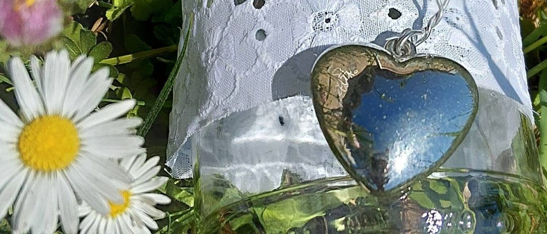 Ein klares Glasglas, geschmückt mit einem weißen Spitzenband, ist von Gänseblümchen umgeben. Ein herzförmiges Anhänger mit einer Silberkette hängt im Inneren des Glases.