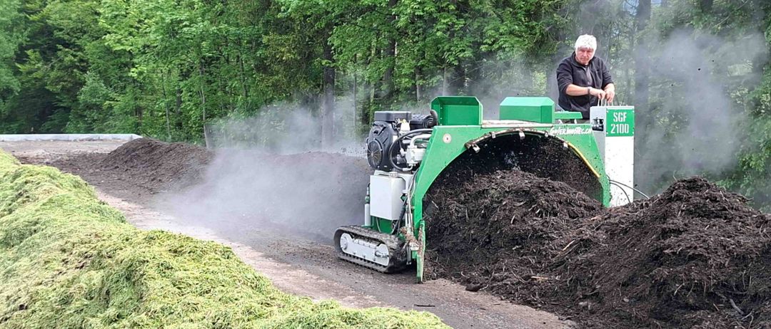 Ein Mann bedient eine grüne Maschine auf einer Straße und sprüht Erdstaub in die Luft. Die Maschine hat einen großen Eimer vorne.