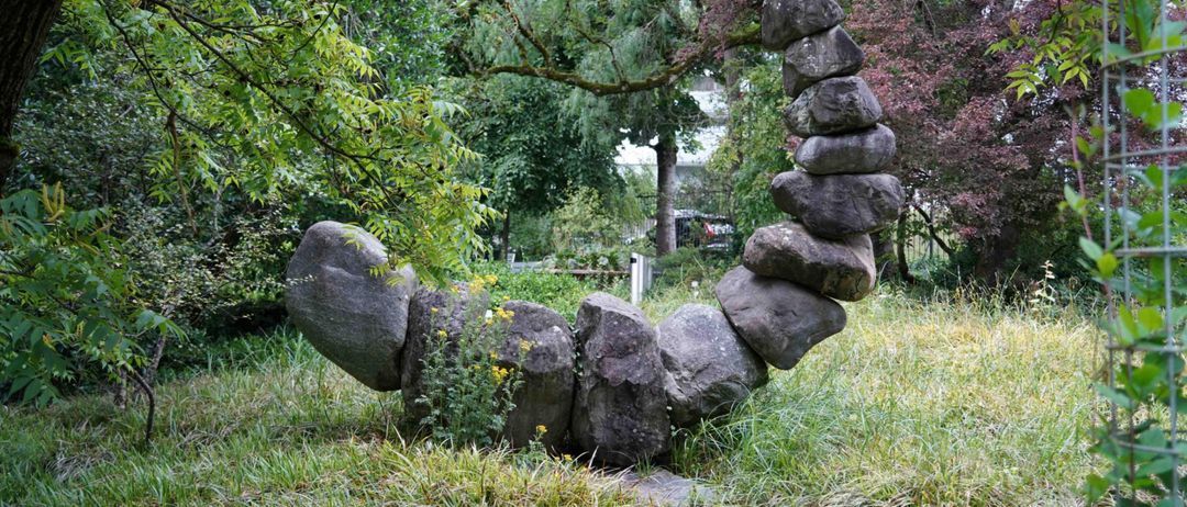 Eine Steinskulptur, die einer Welle oder einer Schlange ähnelt, befindet sich in einem Park mit hohen Bäumen und Gras.