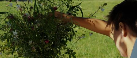 Eine Person, die einen großen Strauß Wildblumen in einem Grasfeld mit Blick auf entfernte Berge und Bäume hält.