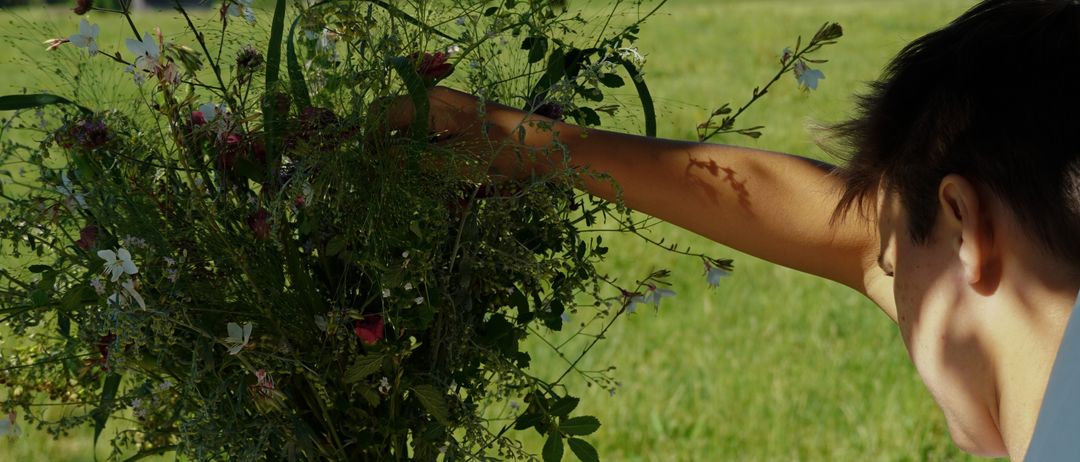 Eine Person, die einen großen Strauß Wildblumen in einem Grasfeld mit Blick auf entfernte Berge und Bäume hält.