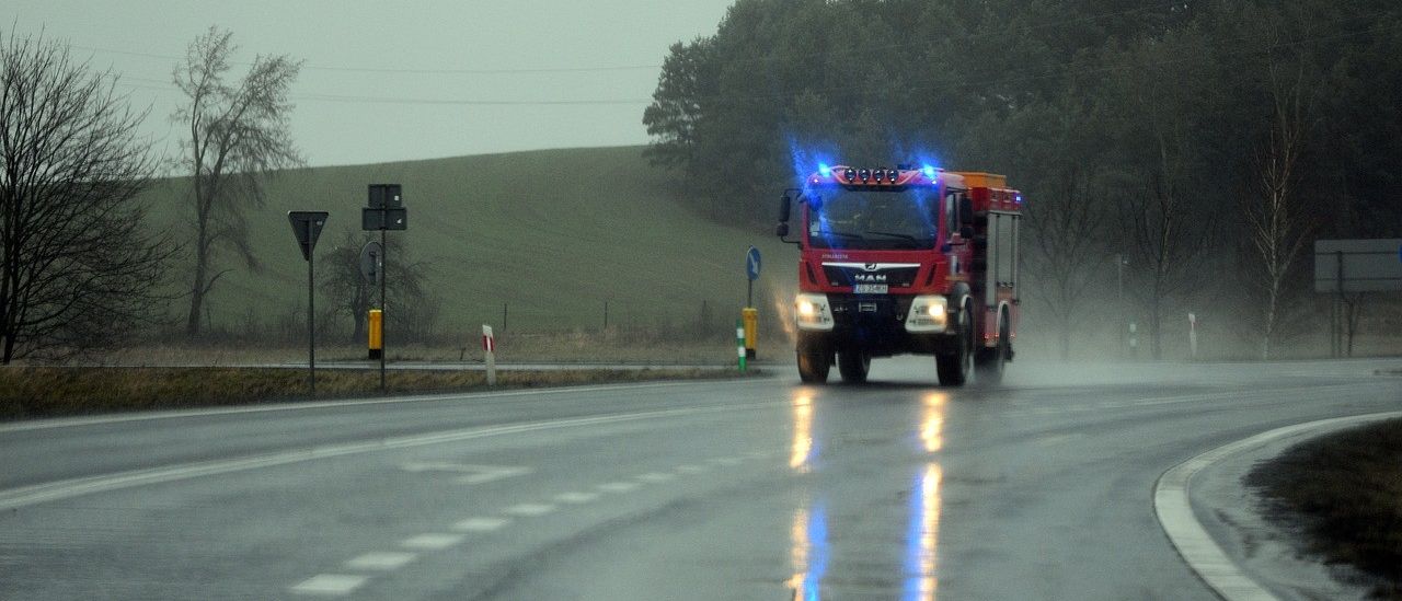 Ein roter Feuerwehrwagen mit blauen Blitzlichtern fährt auf einer nassen Straße mit Bäumen und Hügeln im Hintergrund.