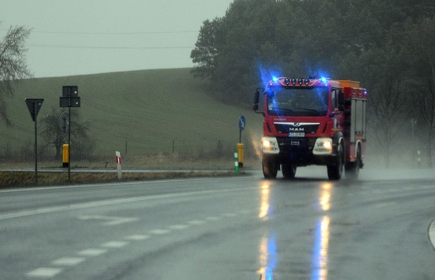 Ein roter Feuerwehrwagen mit blauen Blitzlichtern fährt auf einer nassen Straße mit Bäumen und Hügeln im Hintergrund.