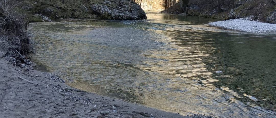 Ein ruhiger Fluss mit einer Brücke darüber, umgeben von Bäumen und einem felsigen Hang. Das Wasser ist ruhig und klar.