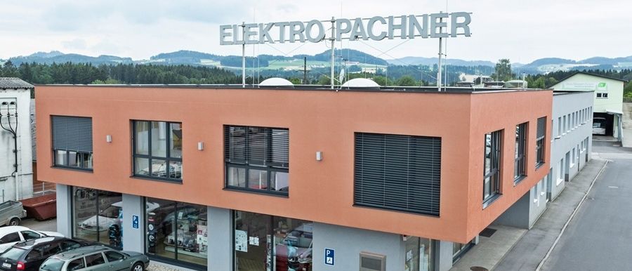 Ein Bild des Bürogebäudes von Elektro Pachner mit geparkten Autos davor. Das Gebäude hat orange Wände und Glasfenster. Es sind zwei Autos davor geparkt.