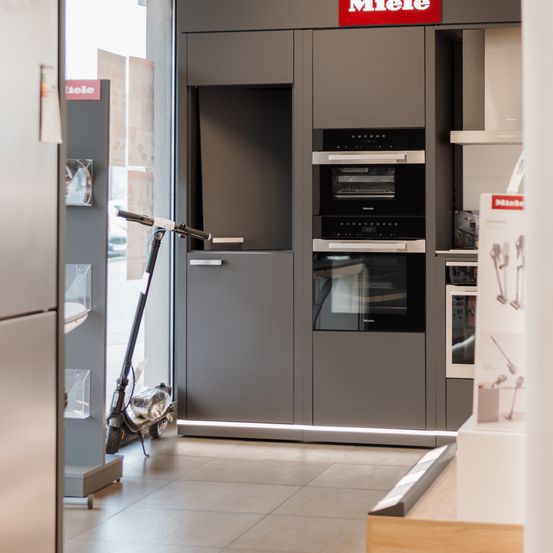 In einem Miele-Geräte-Showroom steht ein Roller neben einem Set eingebauter grauer Schränke. Die Schränke haben ein rotes Miele-Schild über ihnen und enthalten Doppelöfen.