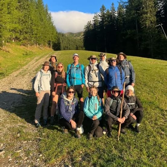 Eine Gruppe von Wanderern steht auf einem Grasfeld und lächelt für ein Foto, mit Bäumen und einem Berg im Hintergrund.