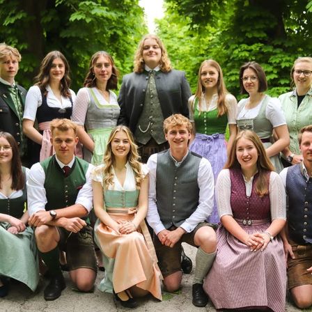 Eine Gruppe von Menschen in traditioneller bayerischer Kleidung posiert für ein Foto in einem Park mit grünen Bäumen und Büschen im Hintergrund.