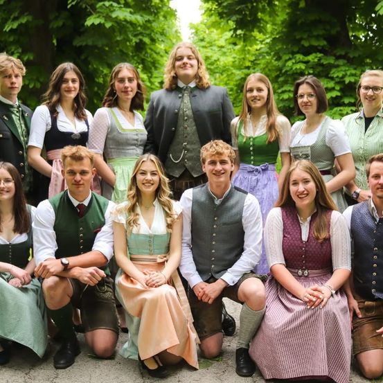 Eine Gruppe von Menschen in traditioneller bayerischer Kleidung posiert für ein Foto in einem Park mit grünen Bäumen und Büschen im Hintergrund.