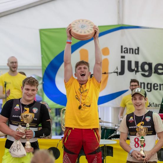 Ein Mann in einem gelben Shirt und roten Hosen hält eine Käsescheibe als Trophäe über seinen Kopf. Zwei andere Männer in Trikots stehen neben ihm, einer hält eine Trophäe. Ein Banner hinter ihnen sagt 'land jugenden'.