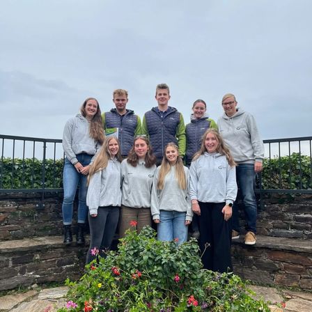 Eine Gruppe von Menschen steht auf Steinstufen und lächelt für ein Foto. Hinter ihnen ist ein schwarzer Metallzaun und es gibt Pflanzen mit Blumen. Sie tragen alle Hoodies.
