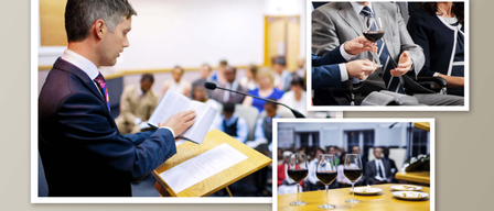 Ein Mann liest von einem Buch an einem Rednerpult, während eine Menge zuschaut. Ein anderer Mann hält ein Weinglas. Weingläser stehen auf einem Tisch mit Essen.