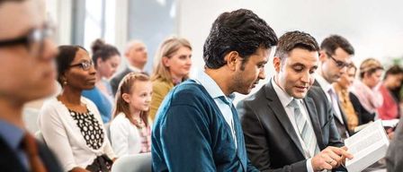 Mehrere Personen sitzen in einem Konferenzraum. Zwei Männer in Anzügen unterhalten sich, einer hält ein Buch. Ein junges Mädchen und eine Frau mit Brille sitzen hinter ihnen.