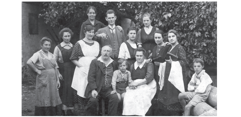 Ein Schwarz-Weiß-Familienfoto zeigt mehrere Generationen vor einem Baum. Ein Mann und eine Frau sitzen mit einem Kind zwischen ihnen, während andere dahinter stehen.