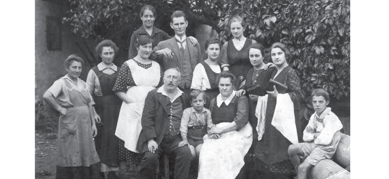 Ein Schwarz-Weiß-Familienfoto zeigt mehrere Generationen vor einem Baum. Ein Mann und eine Frau sitzen mit einem Kind zwischen ihnen, während andere dahinter stehen.