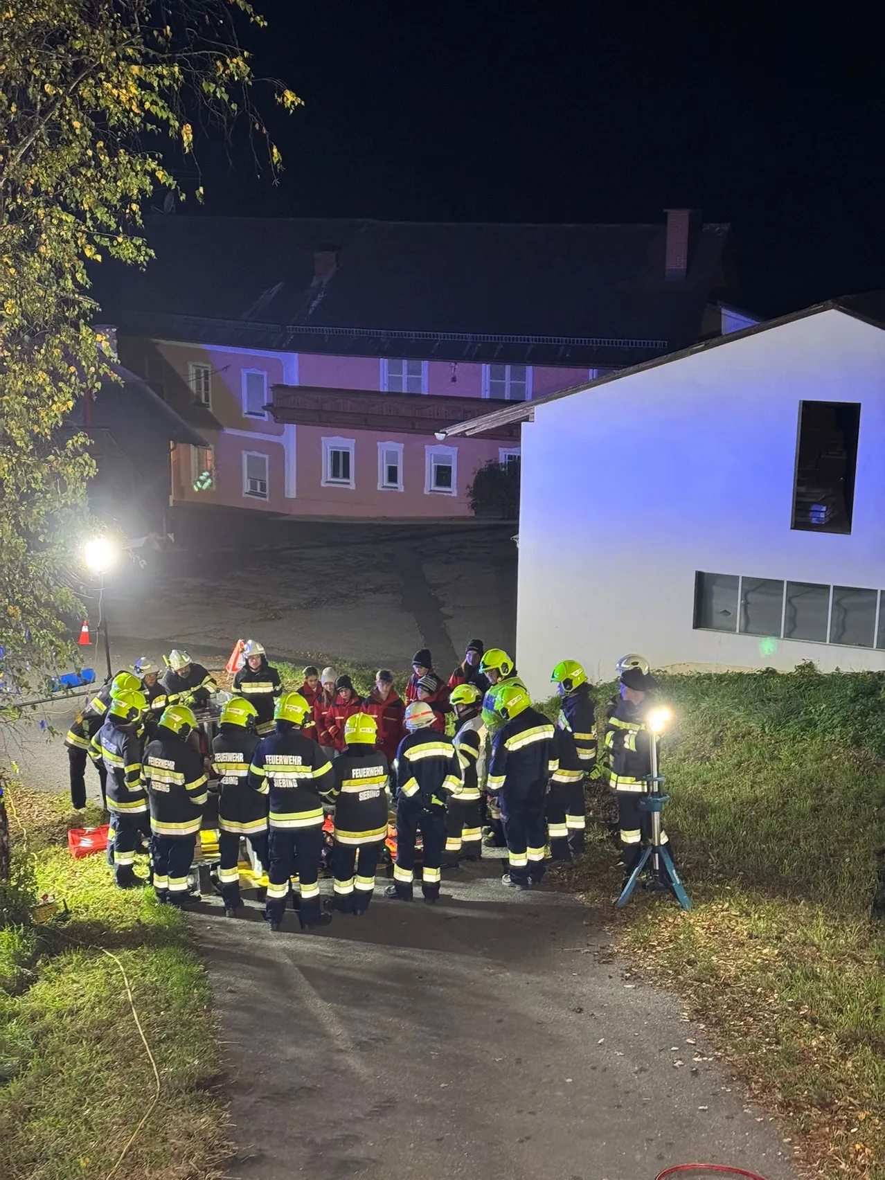Feuerwehrleute in Uniform versammeln sich bei Nacht vor einem Gebäude. Der Bereich ist durch Straßenlaternen beleuchtet und sie stehen auf einem Grasstreifen.