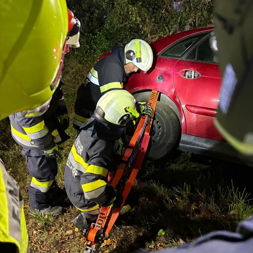 Ein Feuerwehrteam rettet eine Person aus einem roten Auto. Sie benutzen ein Werkzeug, um die Autotür zu entfernen. Die Szene spielt sich draußen bei Nacht ab.