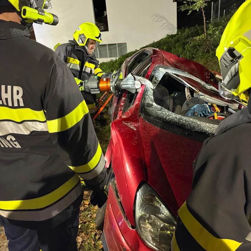 Drei Feuerwehrleute arbeiten an einem roten Auto mit zerbrochener Windschutzscheibe. Einer benutzt ein Werkzeug, um die Scheibe zu schneiden.