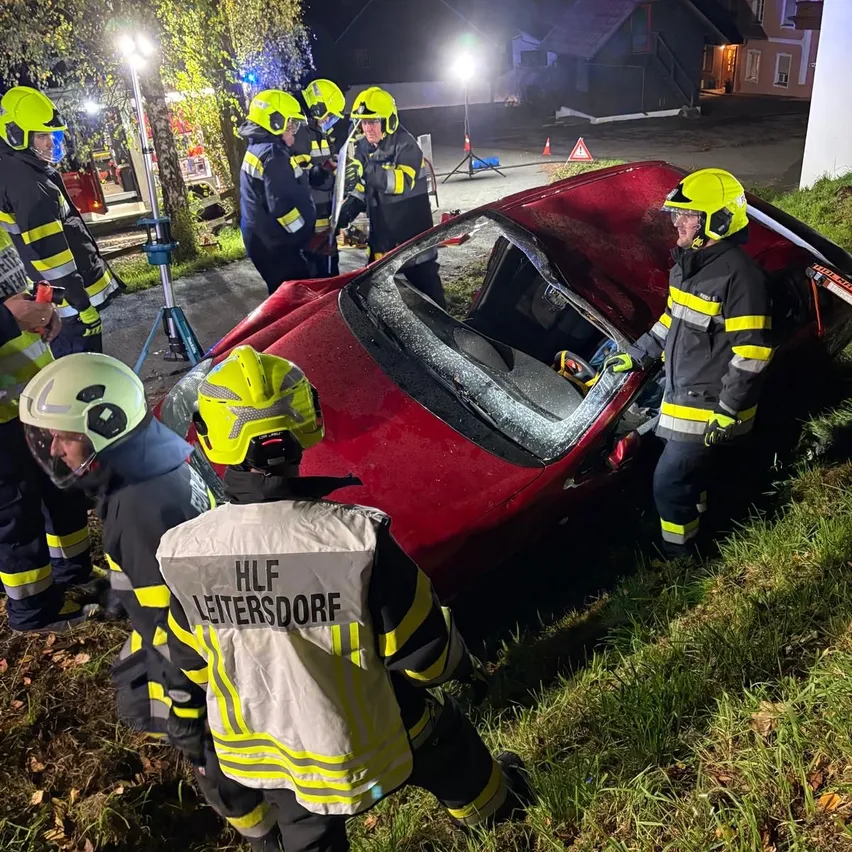 Feuerwehrleute in Schutzkleidung umgeben ein rotes Auto, das nachts auf der Straßenseite zerstört ist.