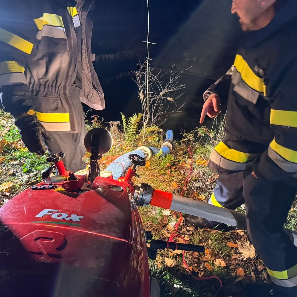 Zwei Feuerwehrleute arbeiten draußen an einer roten Maschine mit der Aufschrift 'Fox'. Einer zeigt auf ein Loch, während der andere einen Schlauch hält. Sie sind von Pflanzen und trockenen Blättern umgeben.