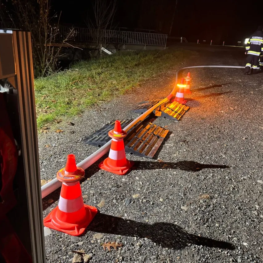 Einsatzkräfte sind bei Nacht draußen mit Verkehrskegel und einer Wasserleitung auf der Straße. Eine Brücke ist im Hintergrund zu sehen.