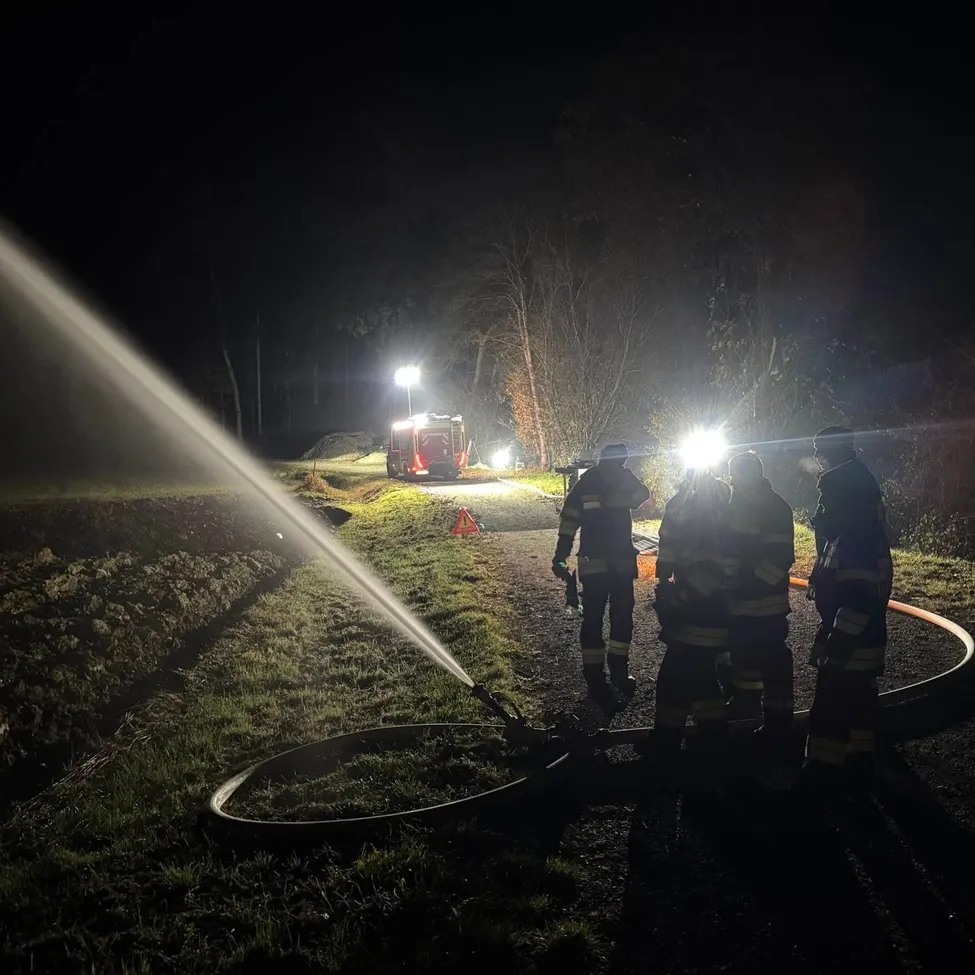 Feuerwehrleute sprühen Wasser aus einem Schlauch auf eine dunkle, nasse Straße bei Nacht. Drei Feuerwehrleute stehen mit einem Feuerwehrwagen und Straßenlaternen im Hintergrund.