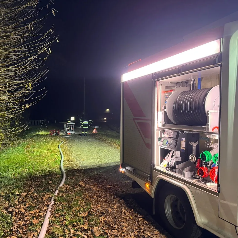 Ein Feuerwehrauto ist nachts am Straßenrand geparkt, die Lichter sind eingeschaltet, Ausrüstung ist sichtbar im Inneren. Zwei Feuerwehrleute stehen in der Nähe.