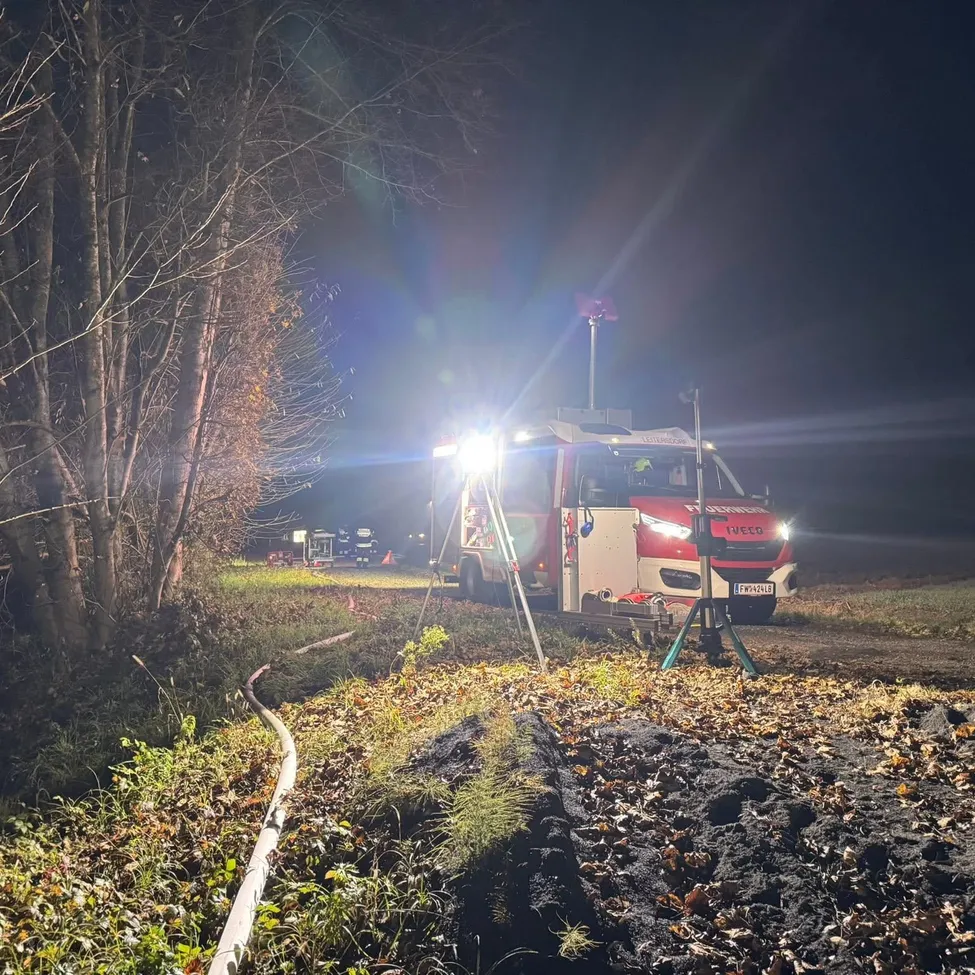 Ein Feuerwehrwagen ist bei Nacht mit eingeschalteten Lichtern am Straßenrand geparkt. Er ist von Bäumen und Gras umgeben.