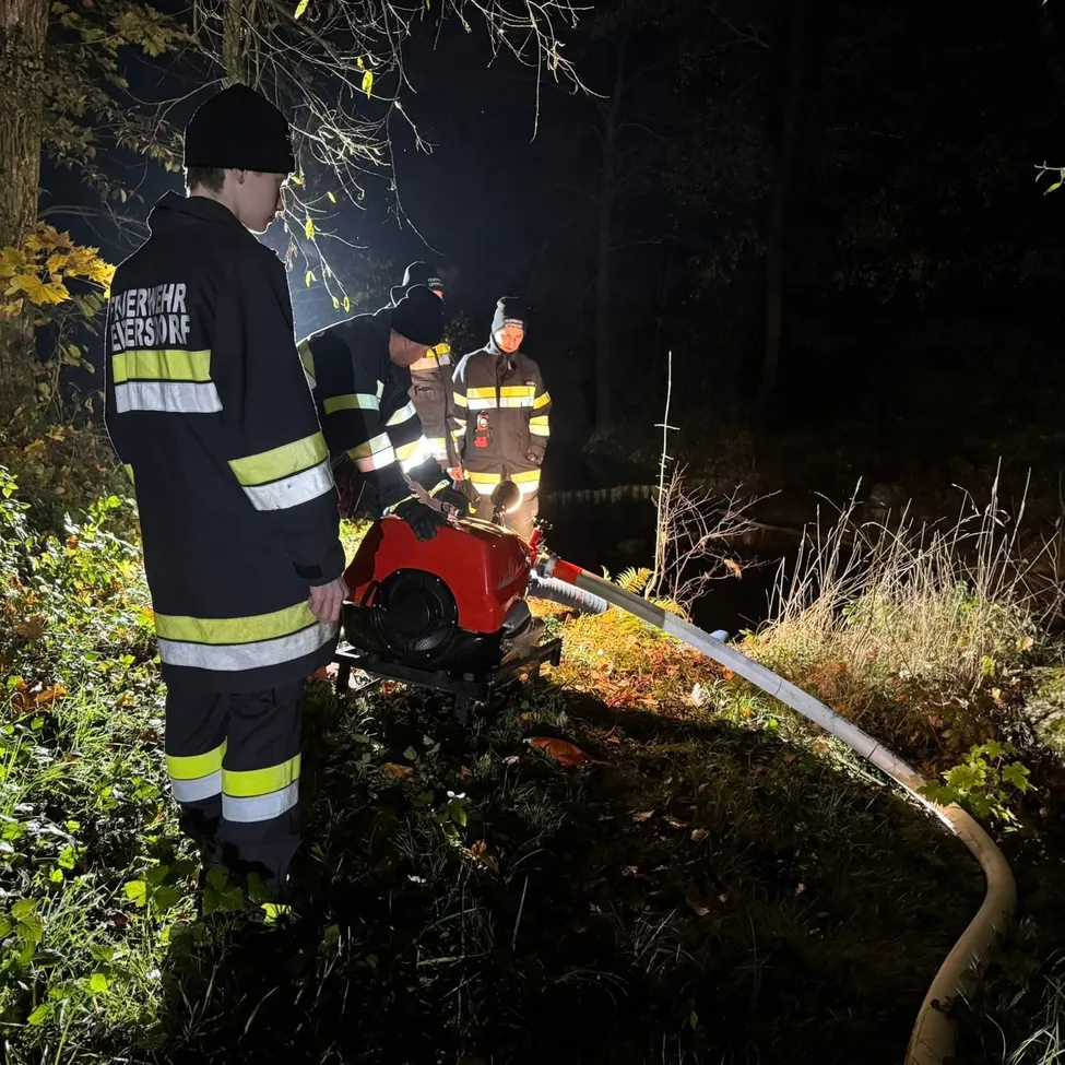 Drei Feuerwehrleute arbeiten nachts an einem Fluss mit einer roten Maschine und einem Schlauch, während einer zur Seite steht.