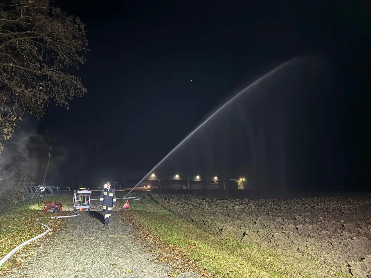 Ein Feuerwehrmann steht bei Nacht auf einem Kiesweg, mit einem Wasserstrahl in der Luft. Ein Gebäude ist in der Ferne sichtbar, und in der Nähe befindet sich ein Verkehrskegel.