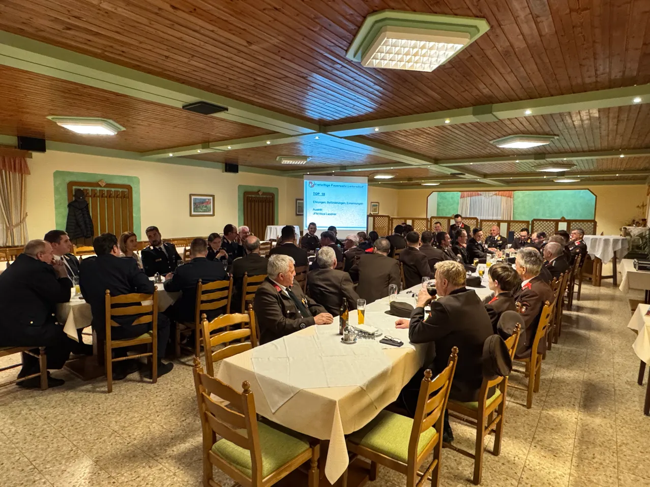 Eine Gruppe von Militäroffizieren sitzt um einen langen Tisch in einem Konferenzraum, wobei eine Projektionsleinwand Informationen dahinter anzeigt.