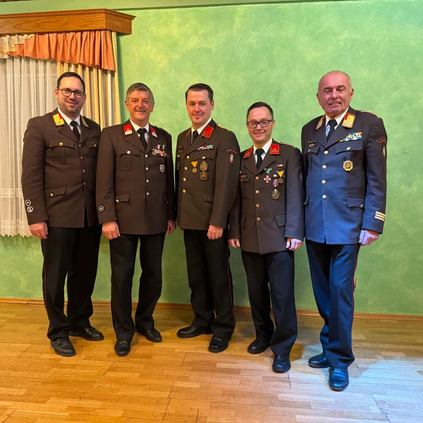 Fünf Männer in Militäruniformen stehen lächelnd nebeneinander vor einer grünen Wand. Sie tragen Medaillen und stehen auf einem Holzboden.
