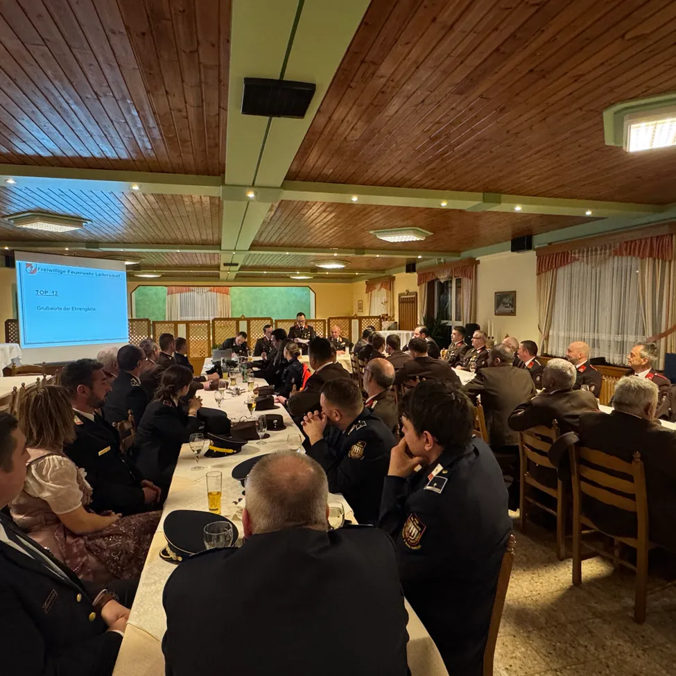 Eine Gruppe von Militärangehörigen und Gästen sitzt um einen langen Tisch in einem Raum mit Holzdecke. Eine Projektionsleinwand befindet sich auf der linken Seite.