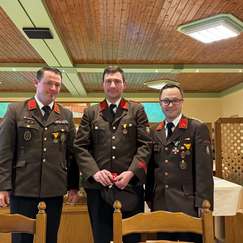 Drei Männer in Militäruniformen stehen nebeneinander in einem Raum. Sie lächeln alle und scheinen für ein Foto zu posieren. Hinter ihnen steht ein hölzerner Tisch mit einem weißen Tuch. Die Decke hat ein hölzerne Design mit Lichtern.