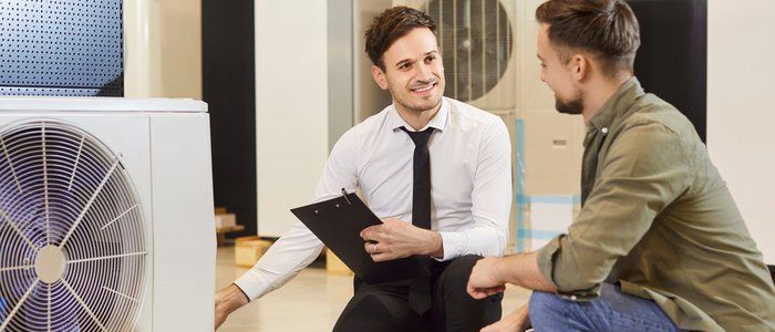 Zwei Männer sind in einem industriellen Umfeld, einer hält eine Clipboard und der andere lächelt. Hinter ihnen stehen weiße Klimaanlagen.
