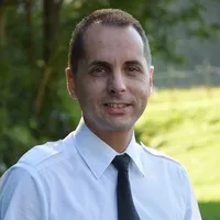 Ein Mann in weißem Hemd und schwarzer Krawatte steht lächelnd im Freien, mit verschwommenem grünen Hintergrund.