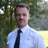 Ein Mann in weißem Hemd und schwarzer Krawatte steht draußen, lächelt und schaut nach rechts, mit einer grasigen Fläche und Bäumen im Hintergrund.