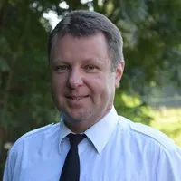 Ein Mann steht im Freien in einem blauen Hemd und einer schwarzen Krawatte, lächelt, mit grünen Bäumen im Hintergrund.
