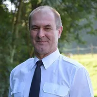 Ein Mann in weißem Hemd und schwarzer Krawatte steht lächelnd vor einem unscharfen grünen Hintergrund.