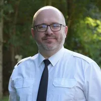 Ein Mann in einem weißen Hemd und einer schwarzen Krawatte steht draußen. Er trägt eine Brille und lächelt. Bäume und Grünpflanzen sind im Hintergrund.