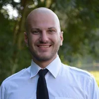 Ein Mann mit kahlem Kopf und weißem Hemd und schwarzer Krawatte lächelt und posiert für ein Foto mit Bäumen im Hintergrund.