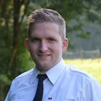Ein Mann in weißem Hemd und schwarzer Krawatte lächelt für ein Foto, mit Bäumen und einem Grasfeld im Hintergrund.