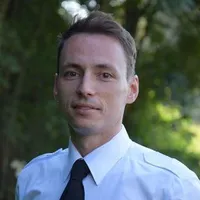 Ein Mann in einem weißen Hemd und schwarzer Krawatte steht vor einem verschwommenen grünen Hintergrund.