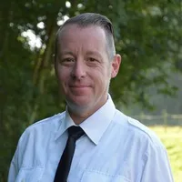 Ein Mann mit einem kurzen Haarschnitt und einem weißen Hemd steht draußen und lächelt. Er trägt eine schwarze Krawatte. Der Hintergrund ist unscharf mit grünen Bäumen.