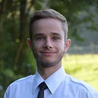 Ein Mann mit kurzen blonden Haaren lächelt und steht draußen. Er trägt ein weißes Hemd und eine schwarze Krawatte. Hinter ihm ist ein unscharfer Hintergrund aus Bäumen und Gras.