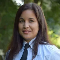 Eine Frau mit langen braunen Haaren lächelt und posiert für ein Foto. Sie trägt ein blaues Hemd und eine schwarze Krawatte. Der Hintergrund ist unscharf mit grünen Bäumen.