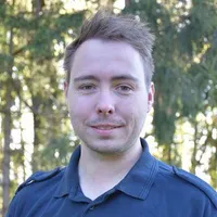 Ein Mann mit kurzen Haaren lächelt und posiert für ein Foto vor einem grünen Hintergrund mit Bäumen. Er trägt ein dunkelblaues Poloshirt.