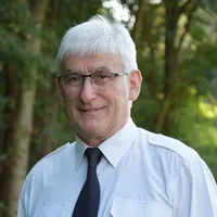 Ein älterer Mann mit weißen Haaren, der eine Brille und ein weißes Hemd mit einer schwarzen Krawatte trägt, steht vor Bäumen.