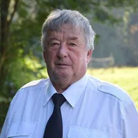 Ein älterer Mann mit grauem Haar steht in einem Park, trägt ein weißes Hemd und eine schwarze Krawatte. Er schaut nach rechts, mit einem unscharfen Hintergrund aus Bäumen und Gras.