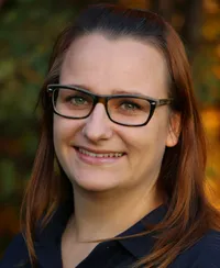 Eine lächelnde Frau mit Brille, langen braunen Haaren und einem blauen Hemd posiert für ein Foto vor einem verschwommenen Herbsthintergrund.